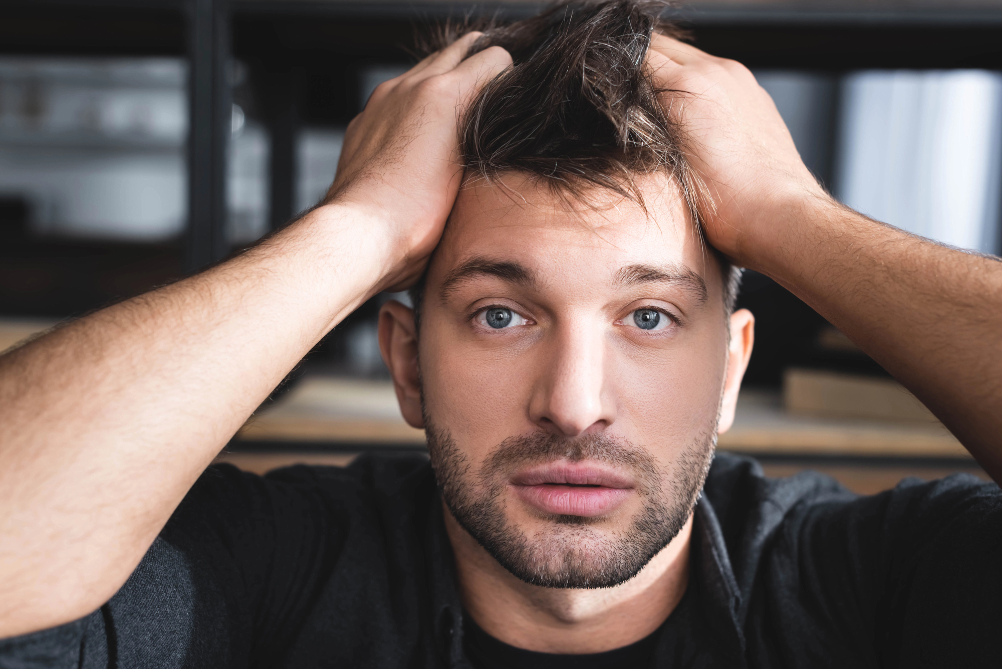 A man with short hair and a beard is looking directly into the camera with his hands on his head, appearing thoughtful or stressed. He is wearing a dark shirt, and the background is blurred, suggesting an indoor setting.