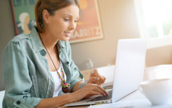 A woman wearing a green shirt and colorful necklace is sitting at a table, using a laptop. Sunlight filters in through a window. A cup is on the table, and a blurred poster is in the background.