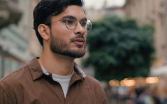 A man wearing glasses and a brown shirt walks down a city street. The background is blurred, showing a tree and buildings. He appears thoughtful and focused as he looks to the side.