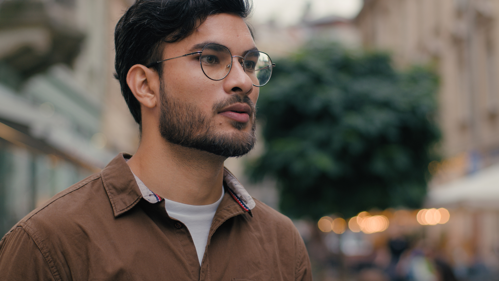 A man wearing glasses and a brown shirt walks down a city street. The background is blurred, showing a tree and buildings. He appears thoughtful and focused as he looks to the side.