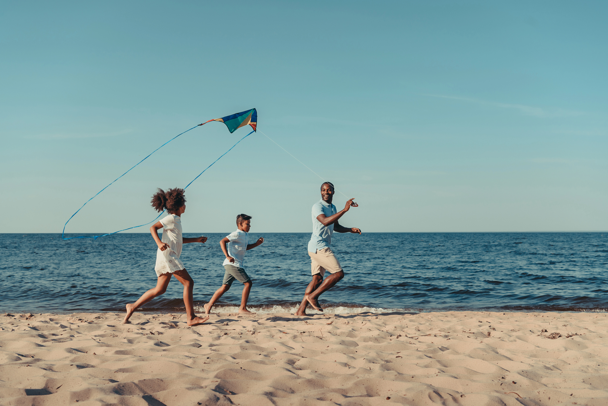 A family of three, wearing casual summer clothes, joyfully runs on a sandy beach while flying a kite. The ocean waves and blue sky provide a serene backdrop.