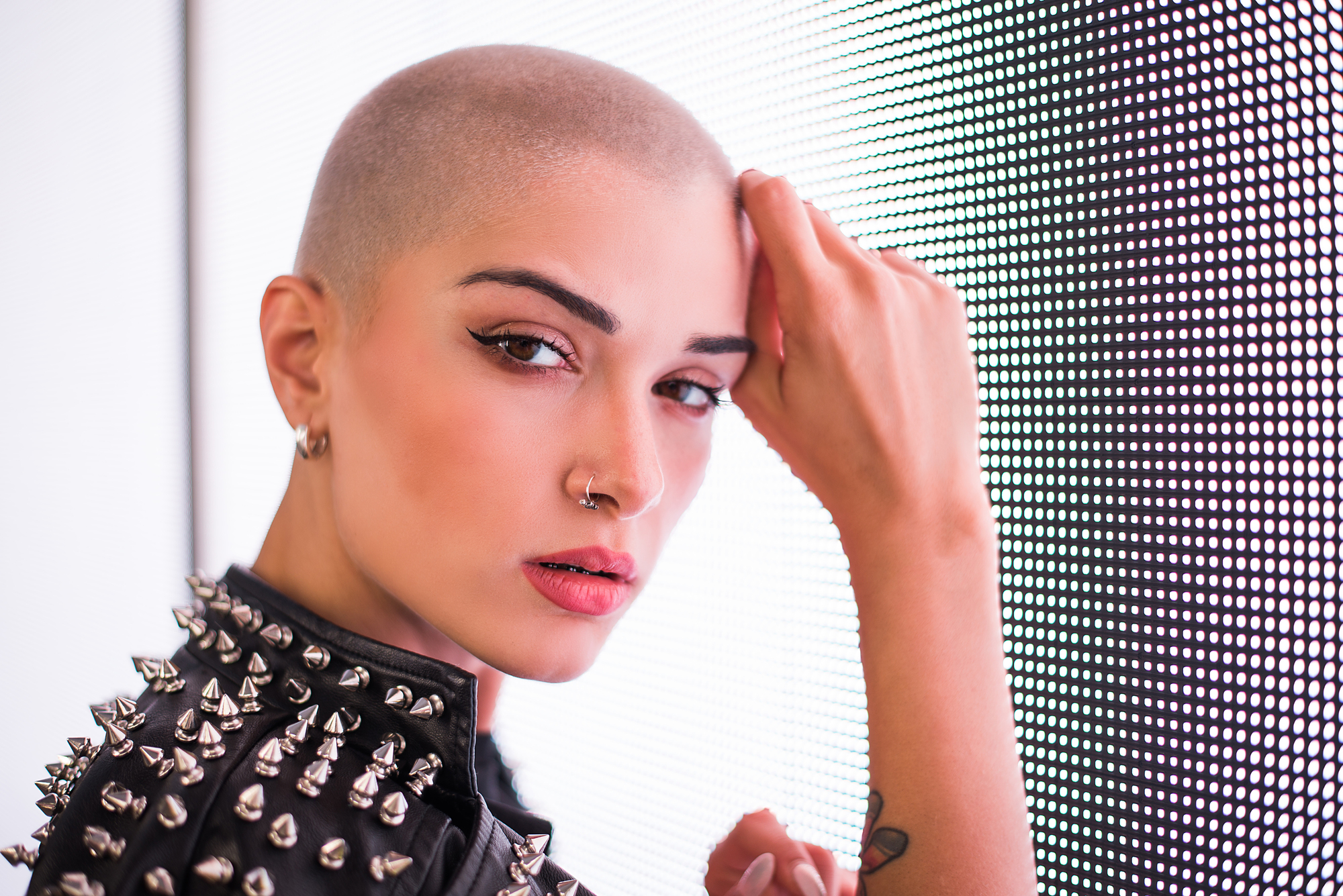 Person with a shaved head and nose ring leans against a lit, dotted background, wearing a studded leather jacket. They have sharp eyeliner and a confident expression.