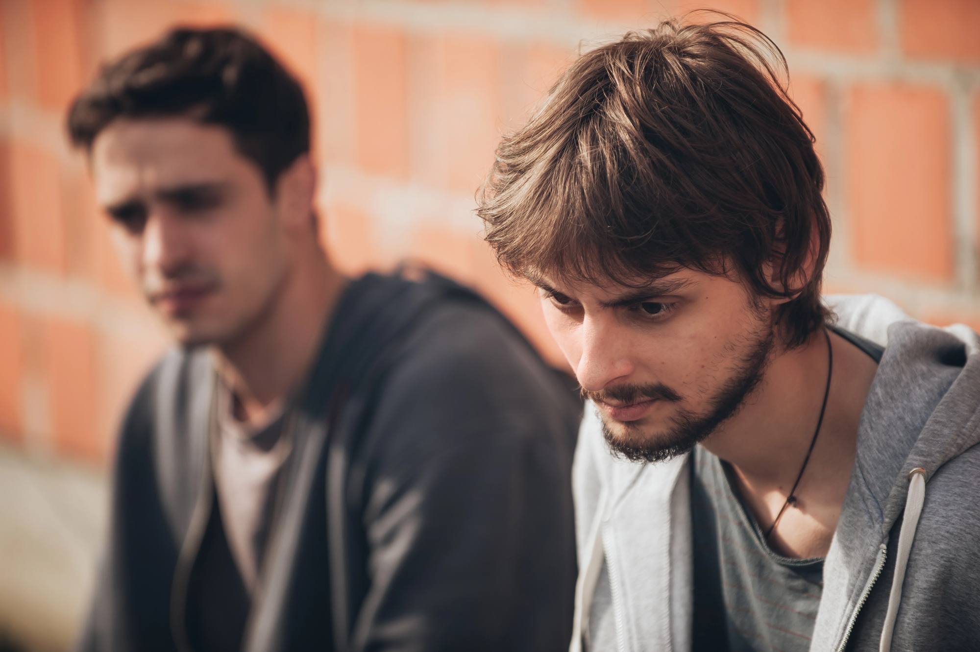 Two young men are sitting outdoors, leaning against a brick wall. The man in the foreground has a pensive expression with slightly tousled hair and a beard. The man in the background is out of focus and appears to be looking in a different direction.