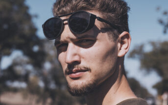 A man with a goatee and short hair stands outdoors, looking at the camera with a serious expression. He is wearing sunglasses pushed up on his forehead. The background is blurred with trees and a clear blue sky.