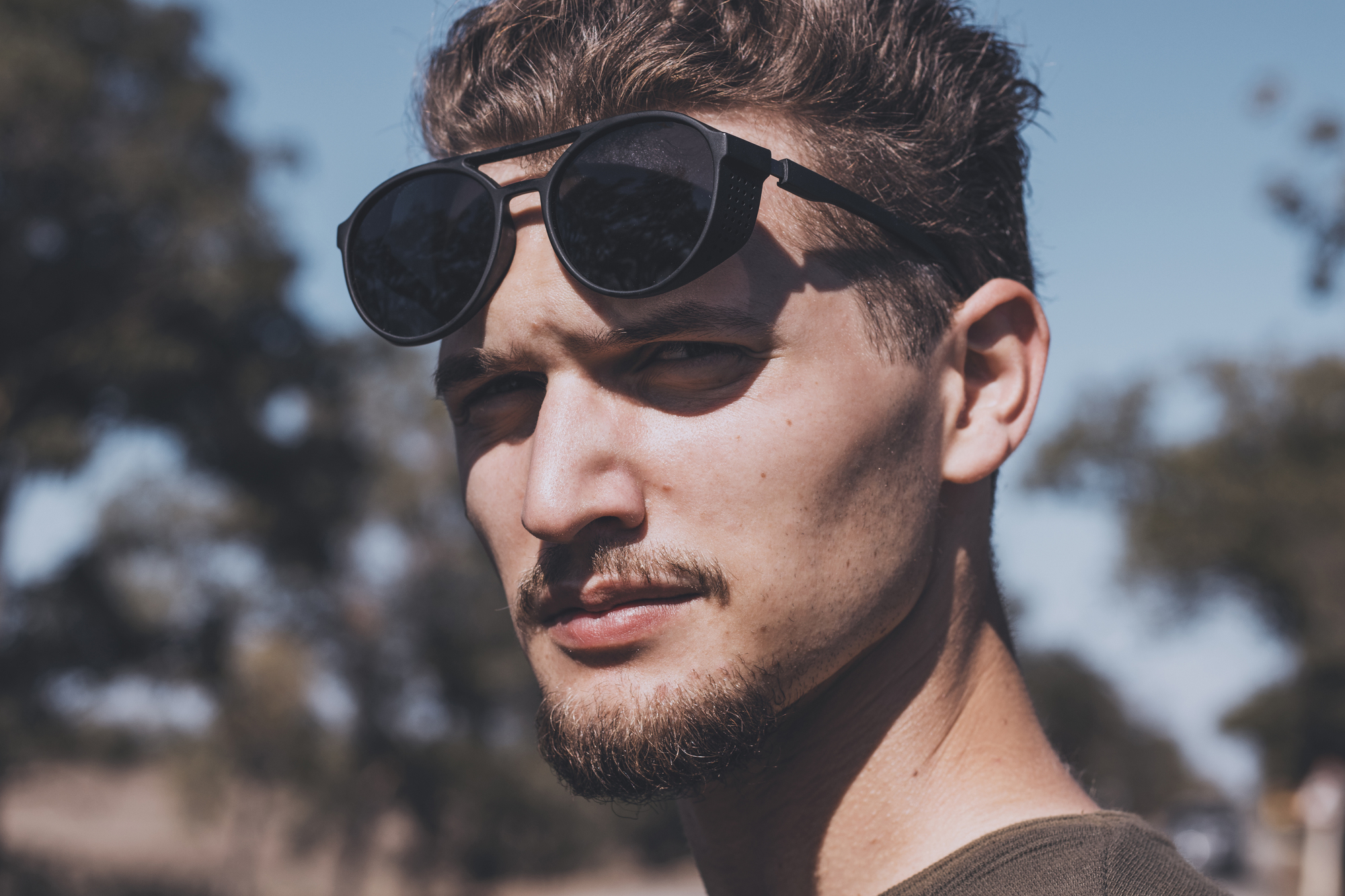 A man with a goatee and short hair stands outdoors, looking at the camera with a serious expression. He is wearing sunglasses pushed up on his forehead. The background is blurred with trees and a clear blue sky.