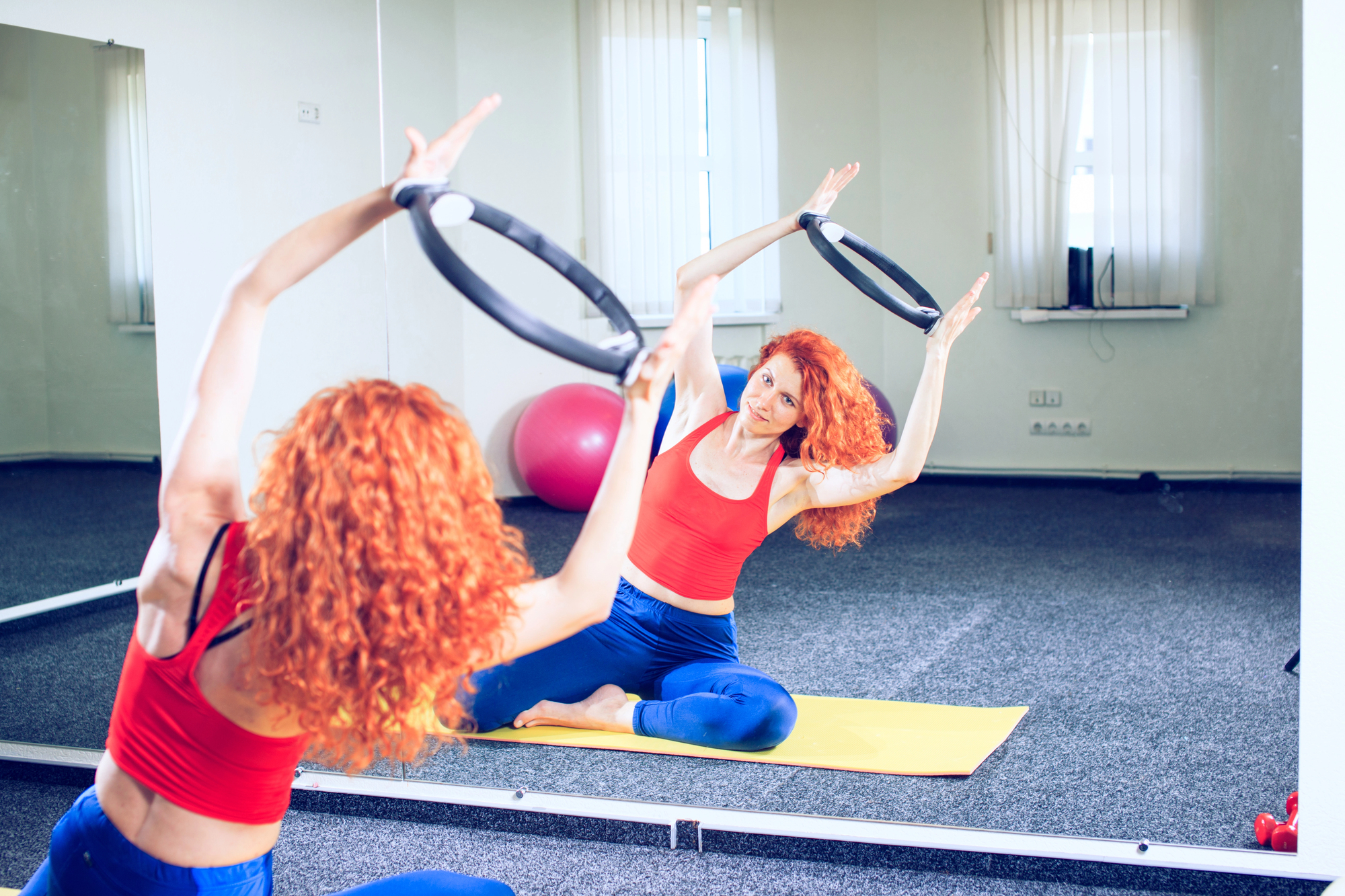 A woman with curly red hair practices yoga in a mirrored studio, sitting on a yellow mat and holding a Pilates ring above her head. She wears a red top and blue pants. Fitness equipment, including a pink exercise ball, is visible in the background.