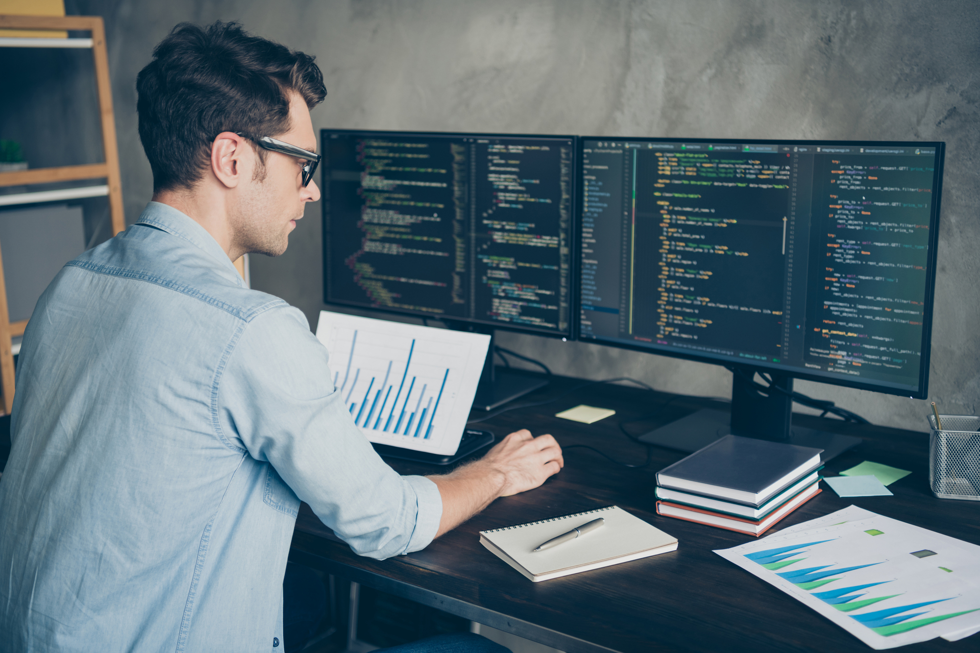 A person wearing glasses is seated at a desk with a computer setup. Two large monitors display lines of code, and a laptop shows a graph. Notebooks and papers are scattered on the desk in a modern office setting.