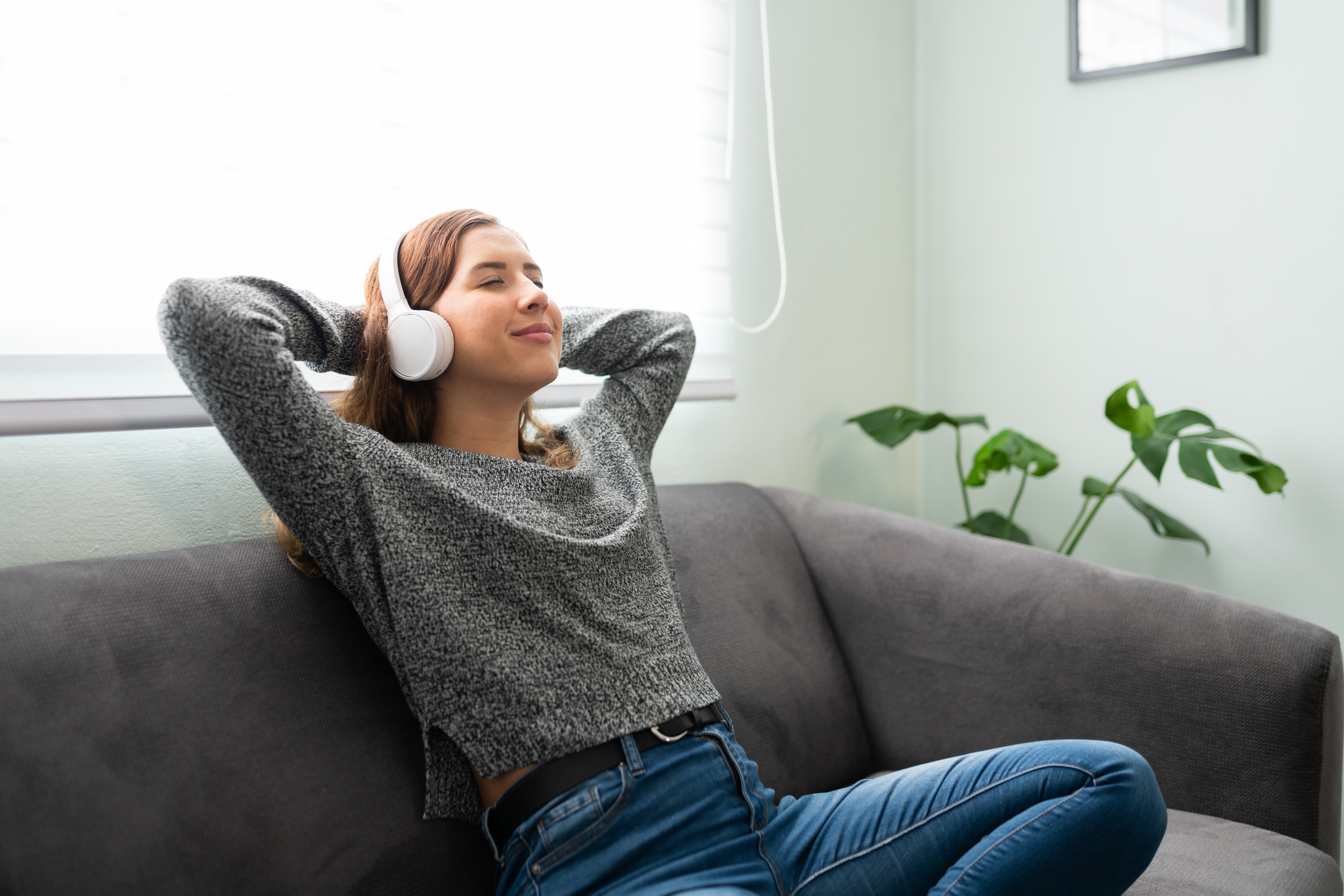 A person wearing headphones relaxes on a gray sofa, smiling with eyes closed and hands behind their head. They are dressed in a gray sweater and jeans. A plant and a window with blinds are visible in the background.