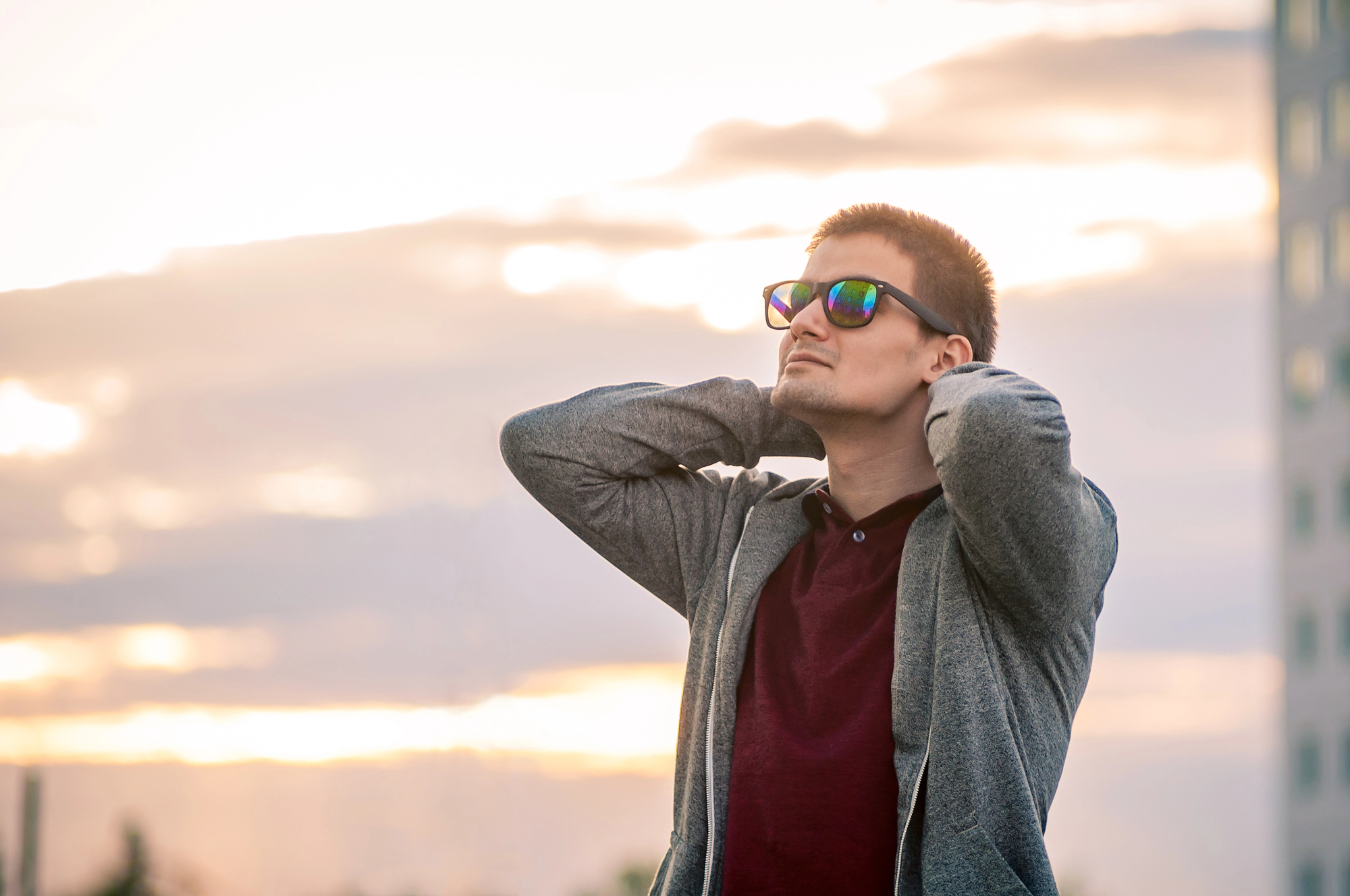 A man wearing sunglasses and a casual outfit stands outdoors with his hands behind his head, looking up. The sky is cloudy with a soft sunset glow in the background.