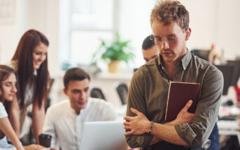 A pensive man with glasses holds a book, standing apart from a group of four people engaged in discussion around a laptop at a workplace. A plant is visible in the background.