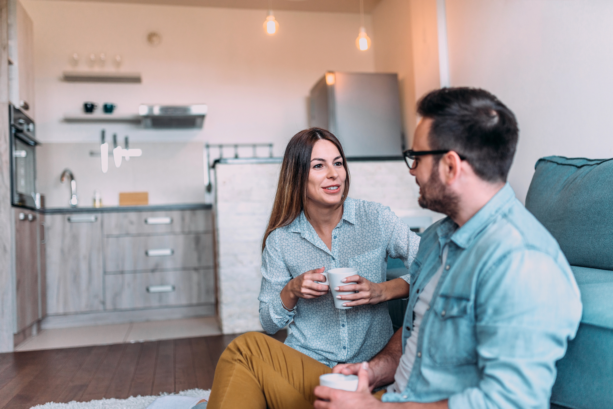 A man and woman sit on the floor in a cozy kitchen, smiling and holding coffee mugs. The kitchen features a modern design with hanging lights and wooden cabinetry. They appear to be engaged in a pleasant conversation.