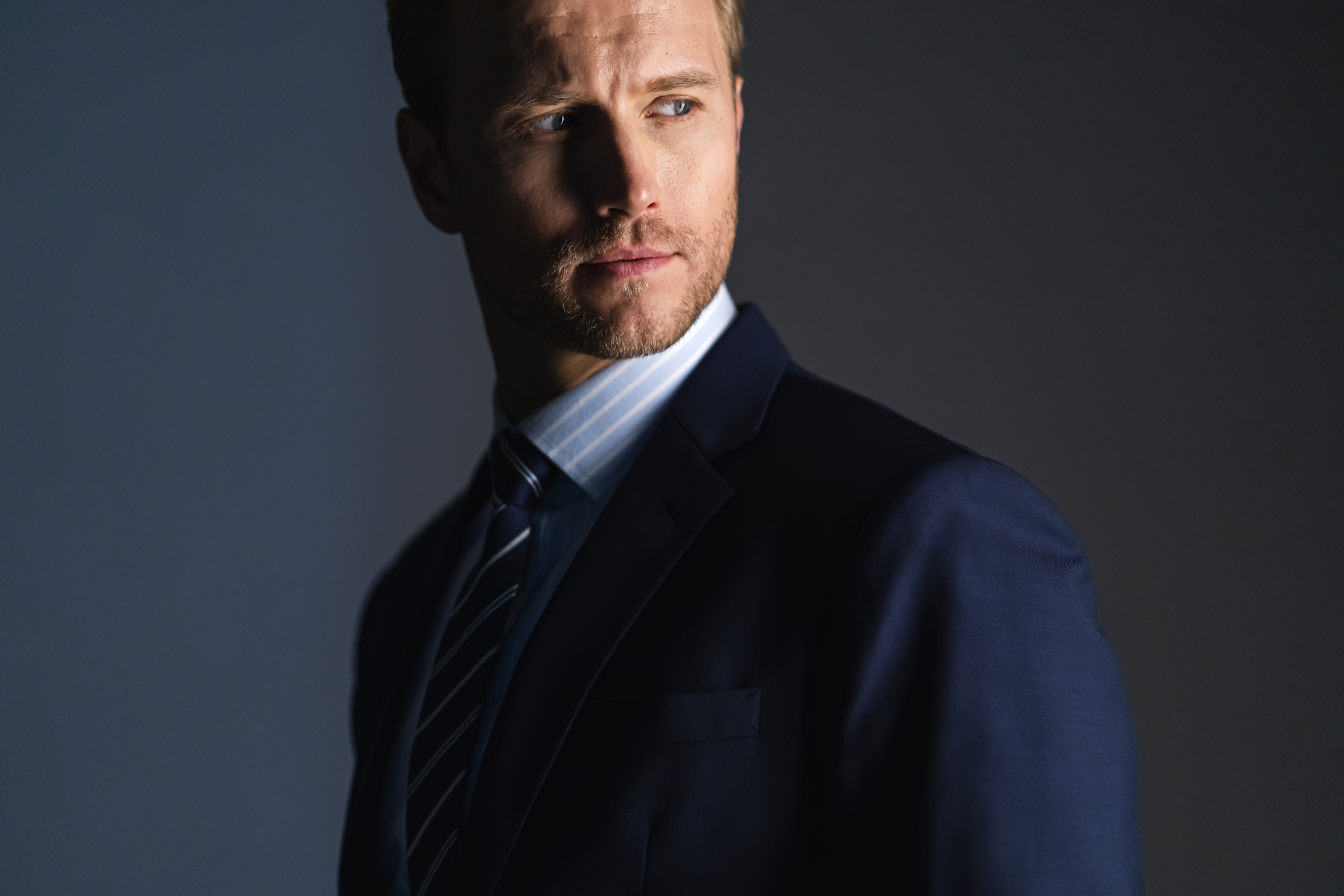 A man in a dark suit and striped tie looks to the side against a dark background, with light casting shadows on his face.