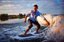 A man in a blue dress shirt and tie surfs on a wave, with his pants rolled up. The sun is setting in the background, casting a warm glow over the scene and the water splashing around him.