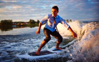 A man in a blue dress shirt and tie surfs on a wave, with his pants rolled up. The sun is setting in the background, casting a warm glow over the scene and the water splashing around him.