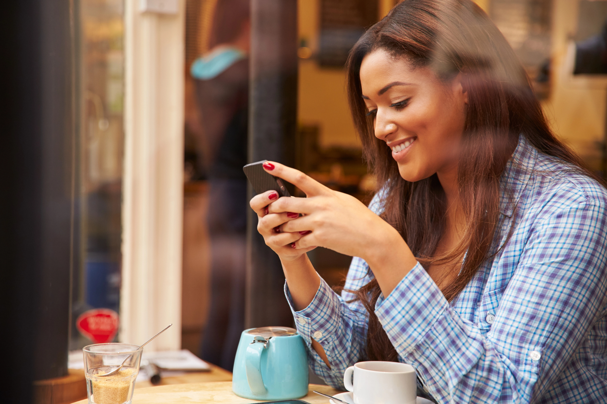 A woman in a plaid shirt sits at a cafe table by a window, smiling as she looks at her smartphone. On the table are a teal teapot, a glass of water, and a white cup. Sunlight filters through the window, creating a warm ambiance.