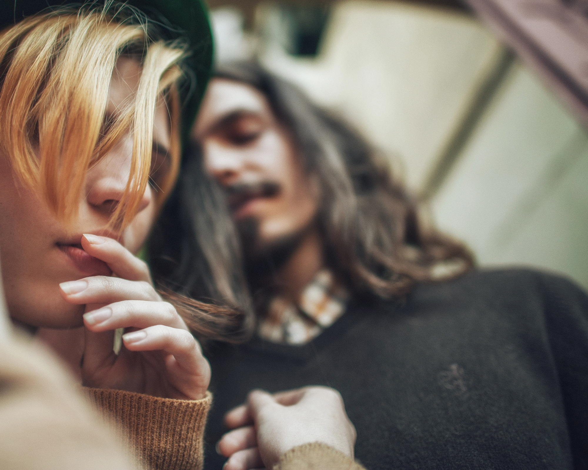 Two people with long hair are close together. One has blonde hair and wears a green hat, while the other has dark hair. The focus is soft, giving a dreamy, intimate feel. They are wearing casual, warm clothing.