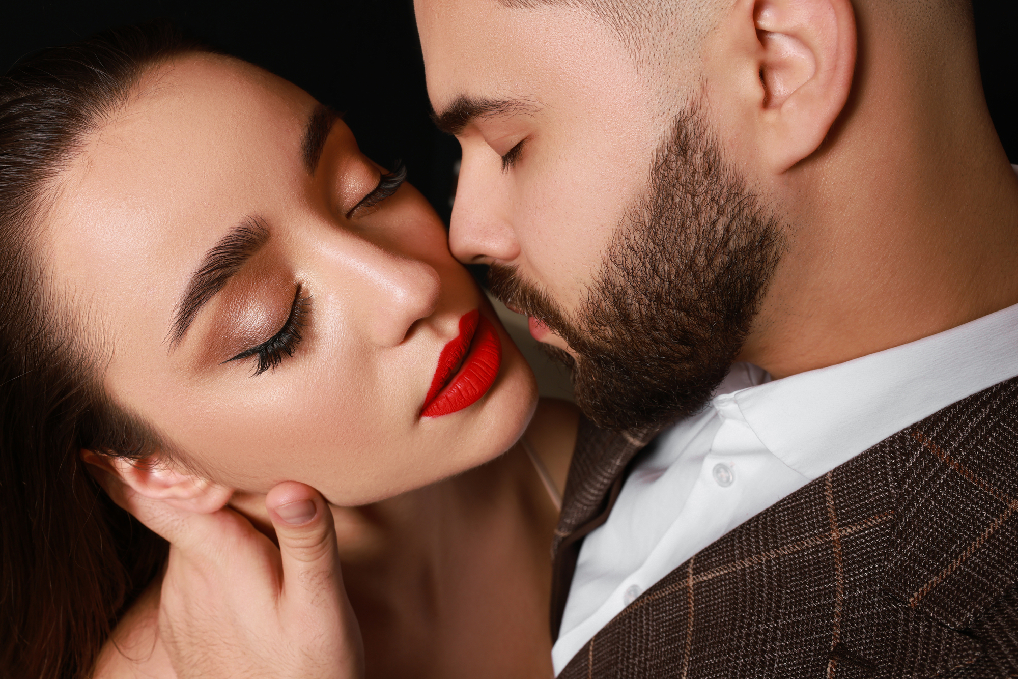 A close-up of a couple about to kiss. The woman has bold red lipstick, winged eyeliner, and long lashes. The man has a beard and is wearing a plaid suit jacket. They are both embracing intimately against a dark background.