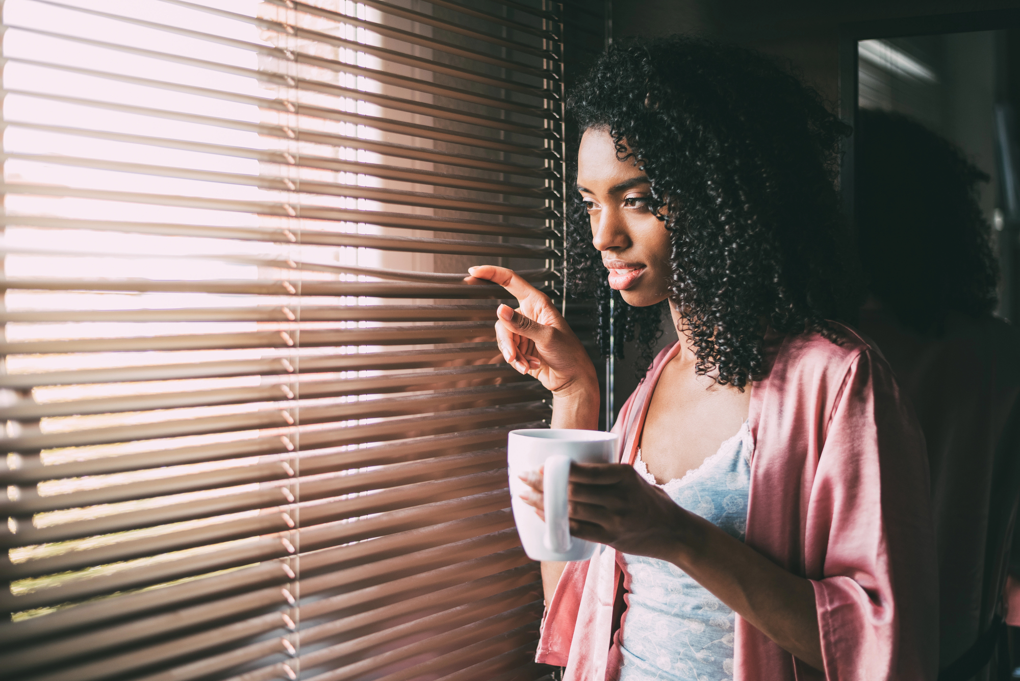 A woman with curly hair, wearing a pink robe, holds a white mug while looking out through blinds. Sunlight filters in, casting soft shadows. She appears thoughtful, standing in a dimly lit room.