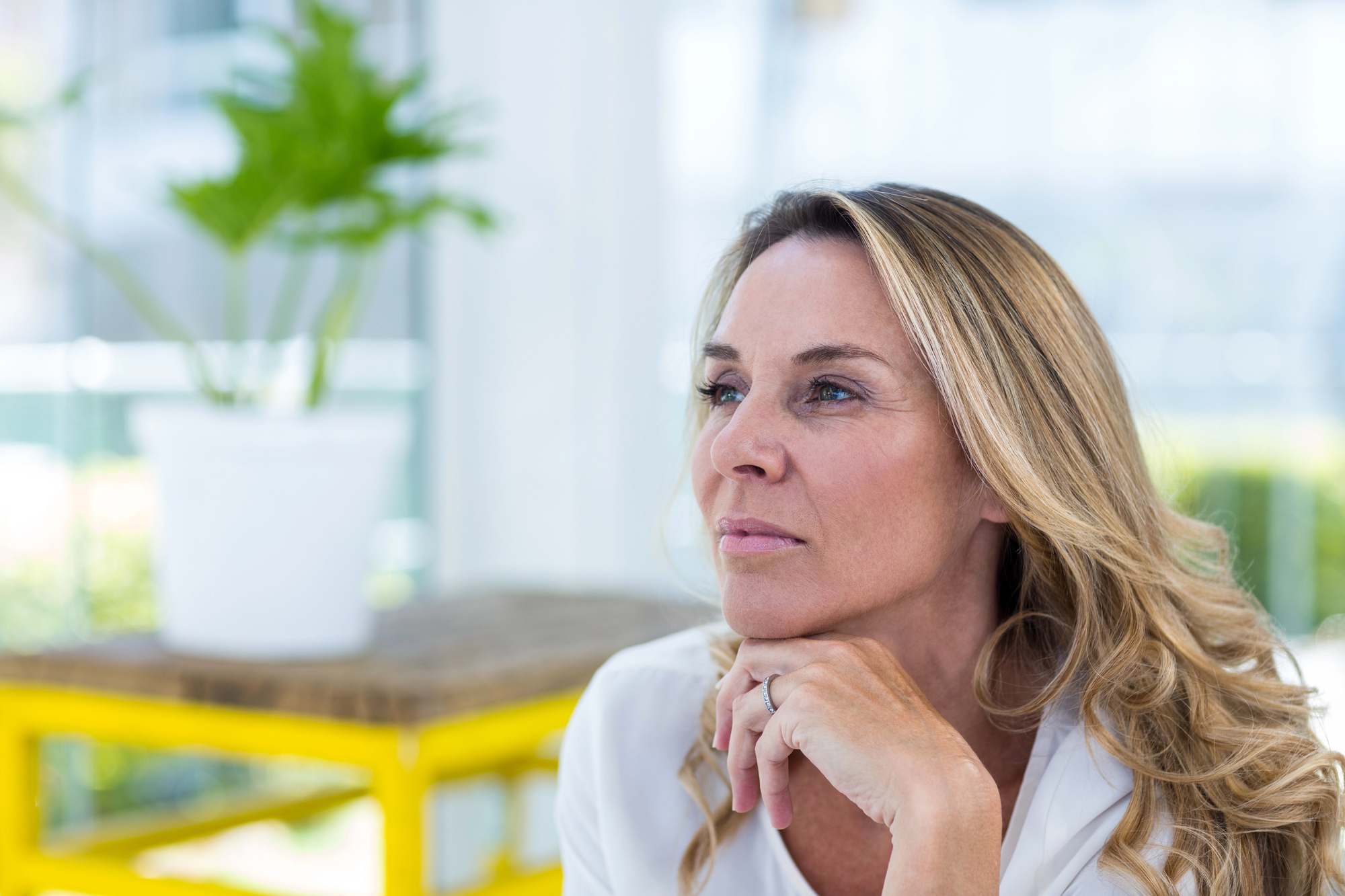 A woman with long blonde hair is sitting indoors thoughtfully, resting her chin on her hand. She is wearing a white top, and there is a potted plant and yellow table in the blurred background.