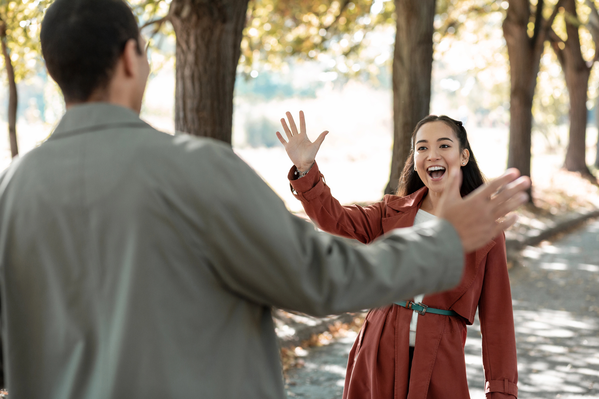 A man and a woman greet each other with open arms and smiles on a sunlit pathway lined with trees. The woman is wearing a red coat, and the man is wearing a gray jacket. The scene conveys a joyful reunion.