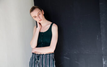 A woman with light skin and blonde hair pulled back, wearing a dark green sleeveless top and black-and-white striped pants, stands against a black and white background. She rests her right hand on her neck and gazes at the camera with a neutral expression.