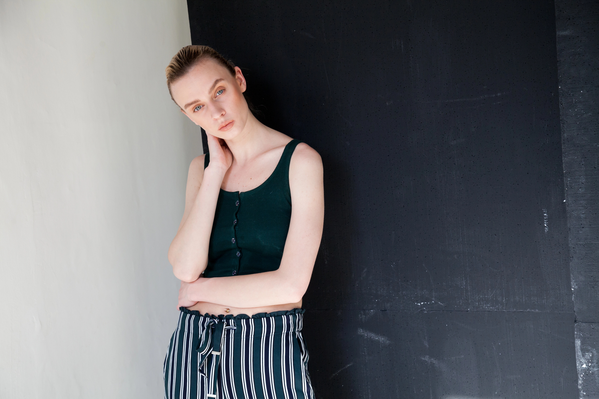 A woman with light skin and blonde hair pulled back, wearing a dark green sleeveless top and black-and-white striped pants, stands against a black and white background. She rests her right hand on her neck and gazes at the camera with a neutral expression.