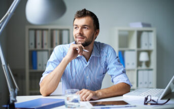 A man in a striped shirt sits at a desk, looking thoughtfully to the side. He holds a pen to his chin, with a laptop, tablet, documents, and a glass of water on the desk. Shelves filled with binders are in the background.