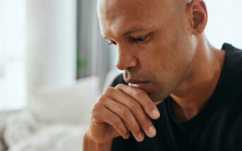 A man with a thoughtful expression rests his chin on his hand, looking down. He is indoors, with a blurred background featuring soft lighting and neutral colors.