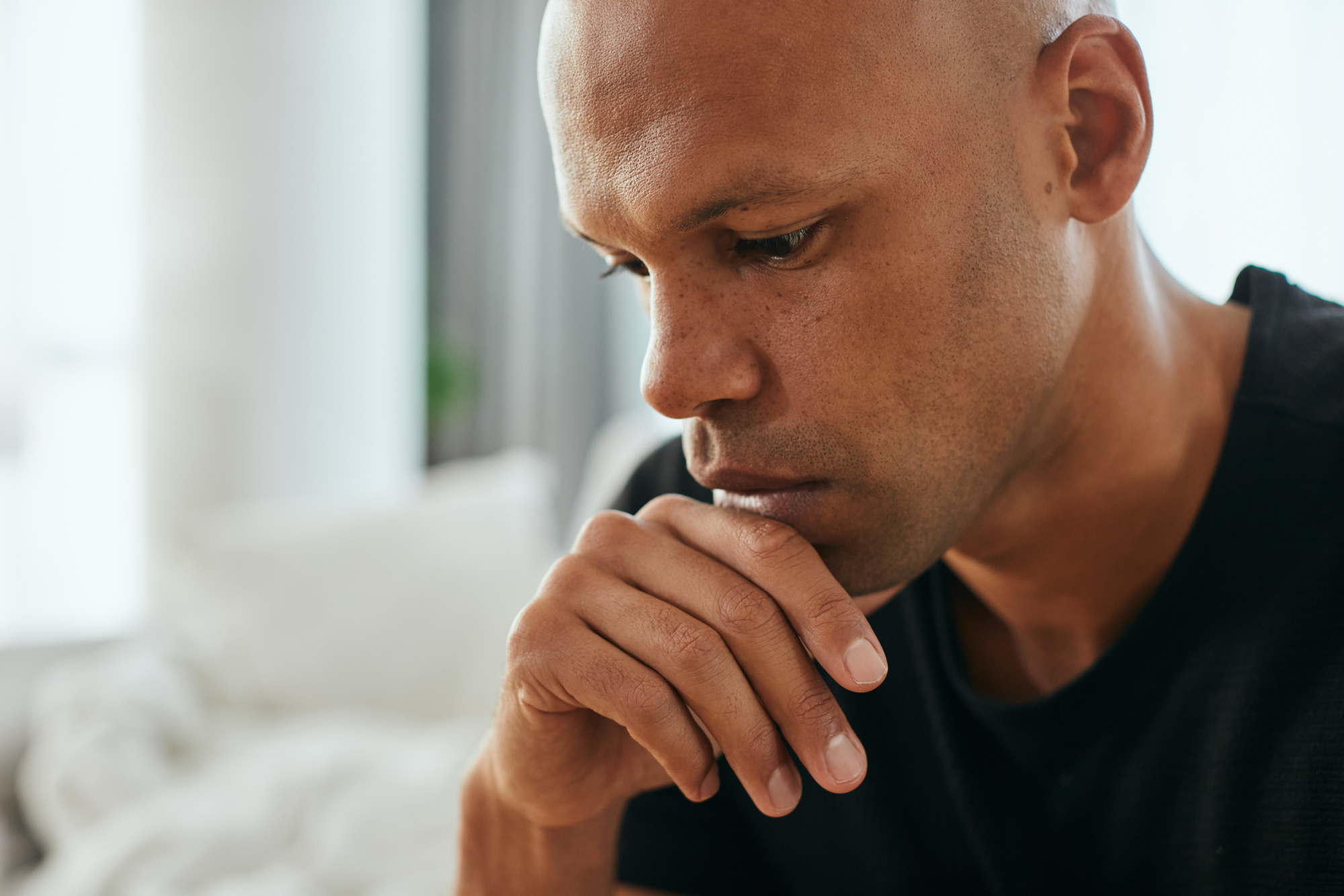 A man with a thoughtful expression rests his chin on his hand, looking down. He is indoors, with a blurred background featuring soft lighting and neutral colors.