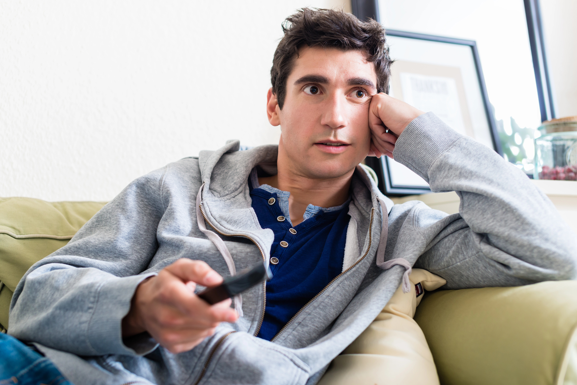 A man in a casual gray hoodie and blue shirt holds a remote control while sitting on a couch. He appears focused, resting his head on one hand, with a framed picture and plant in the background.