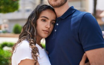 A young woman with long, wavy hair wearing a white shirt leans her head on the chest of a man in a dark blue polo shirt. They are standing close to each other outdoors, with blurred greenery and buildings in the background.