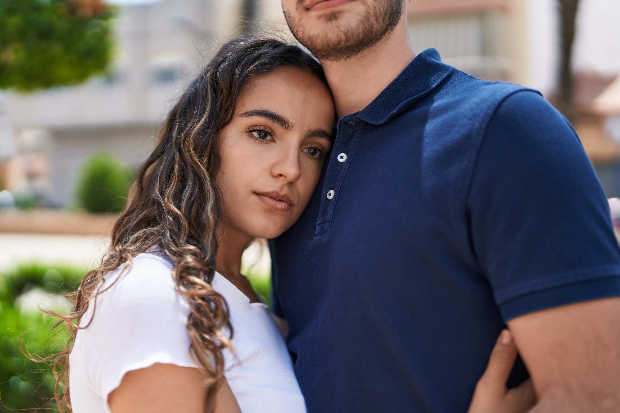 A young woman with long, wavy hair wearing a white shirt leans her head on the chest of a man in a dark blue polo shirt. They are standing close to each other outdoors, with blurred greenery and buildings in the background.