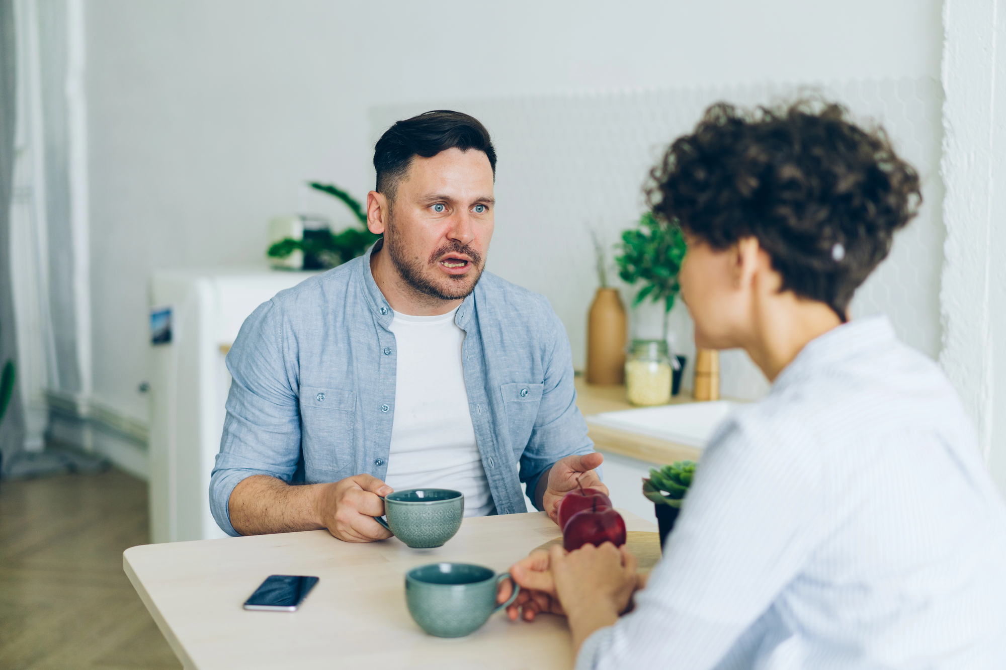 Two people sit at a white table in a modern, minimalistic kitchen, having a conversation. A man with short dark hair holds a mug, wearing a light blue shirt over a white tee. A person with curly hair is seen from the back. A smartphone and décor are on the table.