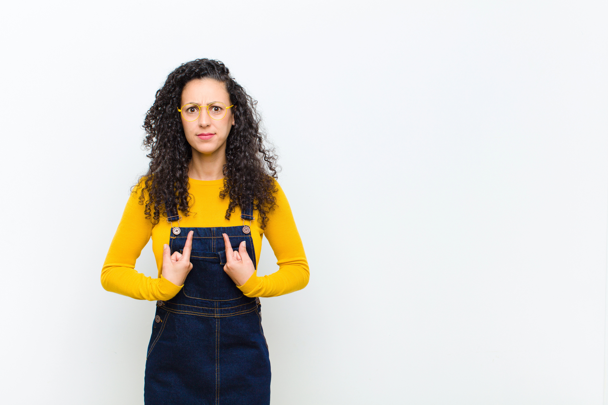 A woman with curly hair and glasses is wearing a yellow long-sleeve shirt and denim overalls. She is standing against a white background, looking at the camera with a puzzled expression, and pointing at herself with both hands.