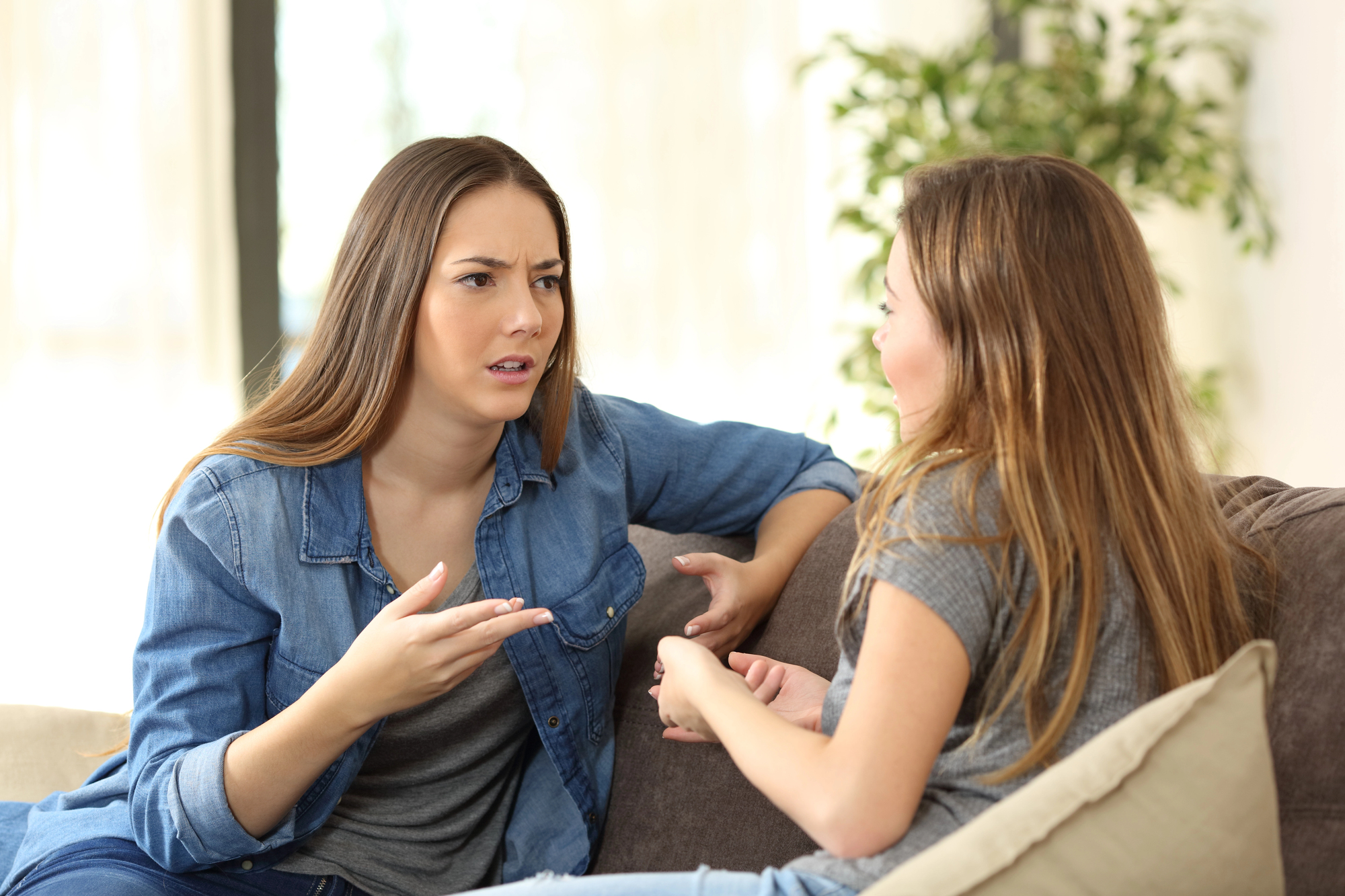 Two women are sitting on a couch. One woman, wearing a denim shirt, appears to be speaking with an expressive gesture, while the other, in a gray shirt, listens attentively. They are in a well-lit room with a large plant in the background.