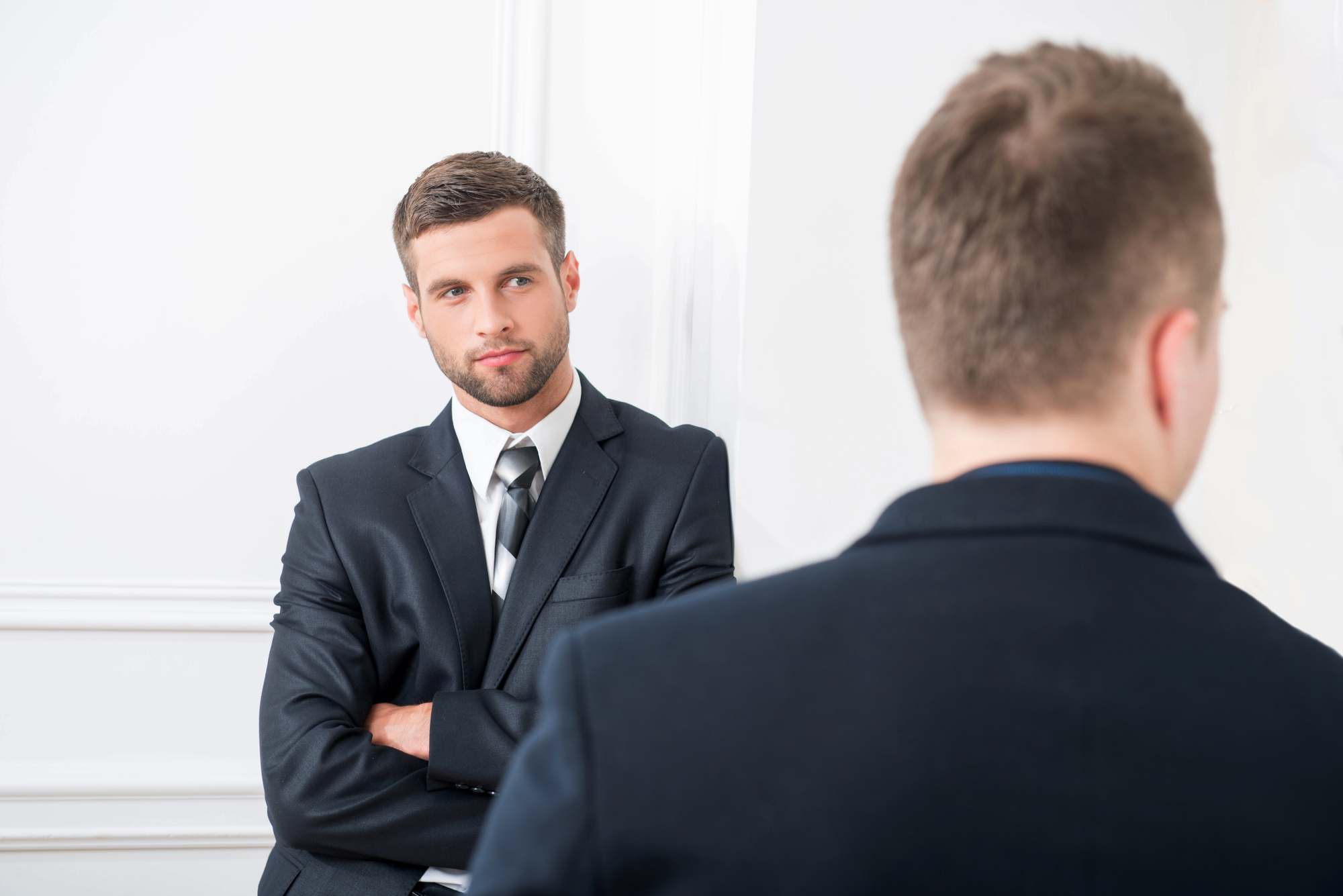 Two men in suits are engaged in conversation in a white room. One man, facing forward, has his arms crossed, while the other has his back to the camera.