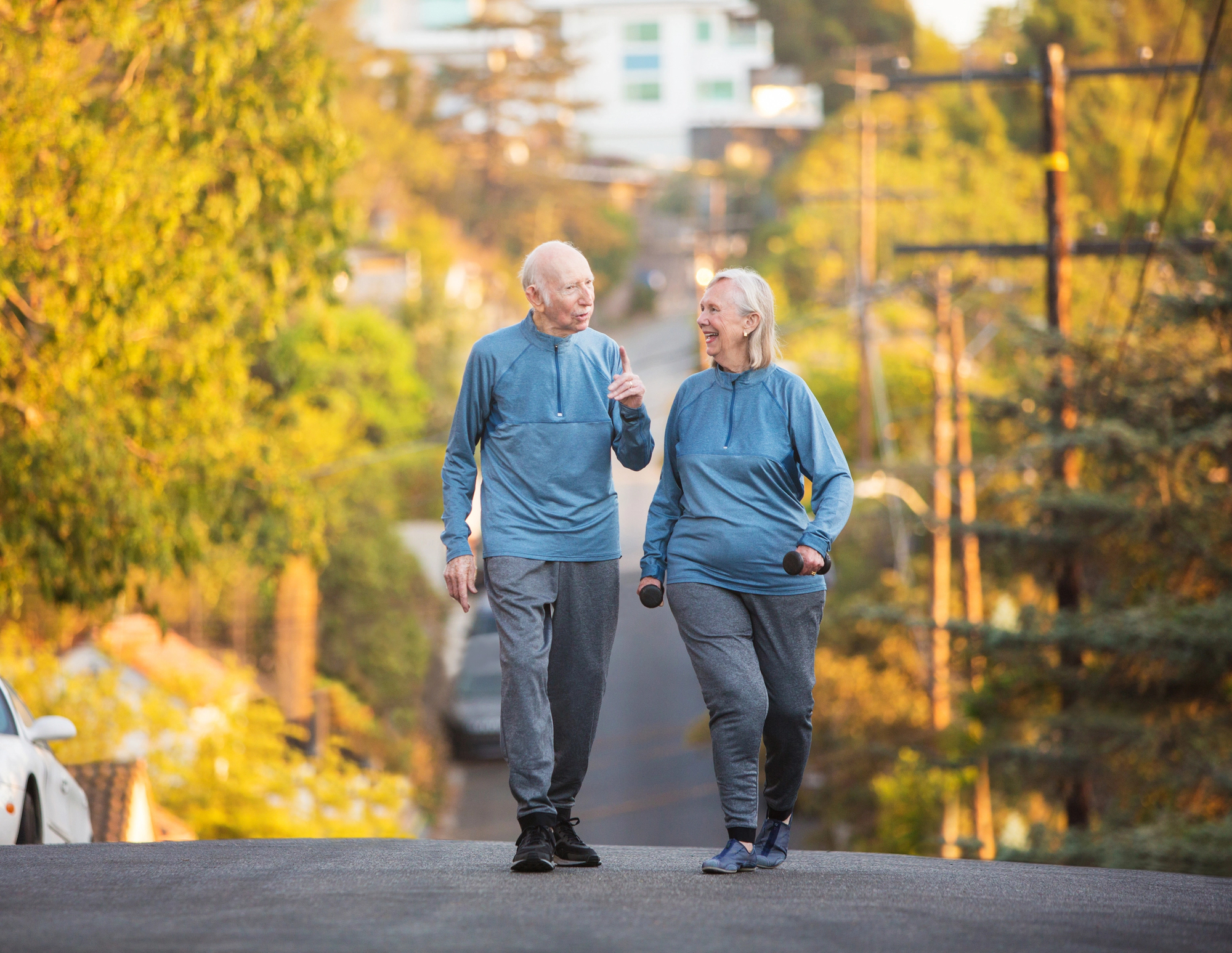 An elderly couple in matching blue and gray activewear walks along a tree-lined street. They smile and chat while holding small dumbbells, enjoying a sunny outdoor exercise together.