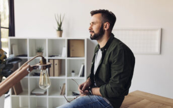 A man with a beard sits on a wooden table, holding glasses in his hands. He looks to the side in a minimalist room featuring white shelves with books and plants, and a vintage-style hanging light bulb nearby.