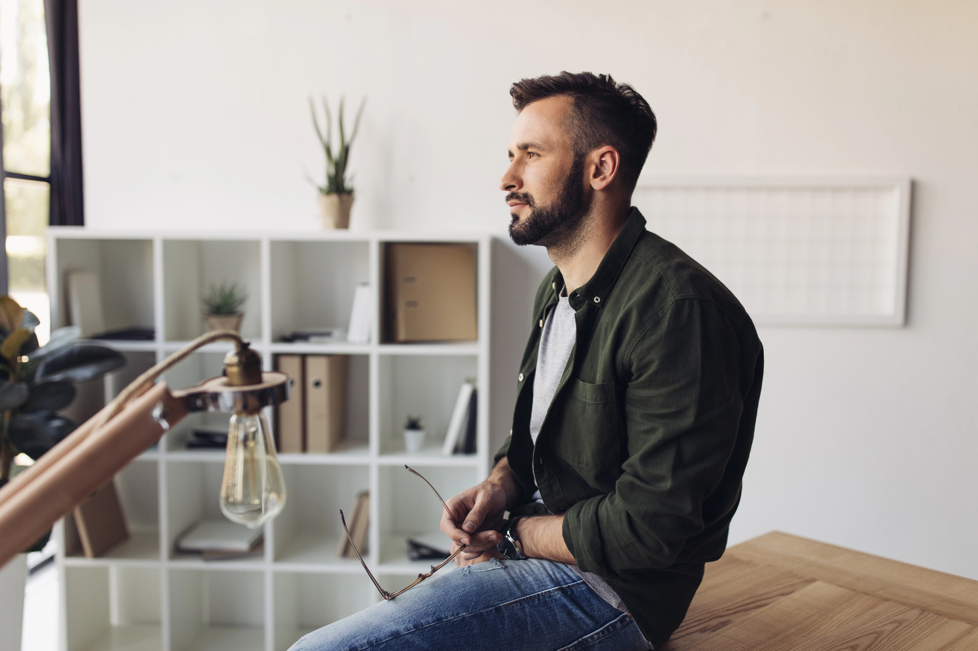 A man with a beard sits on a wooden table, holding glasses in his hands. He looks to the side in a minimalist room featuring white shelves with books and plants, and a vintage-style hanging light bulb nearby.