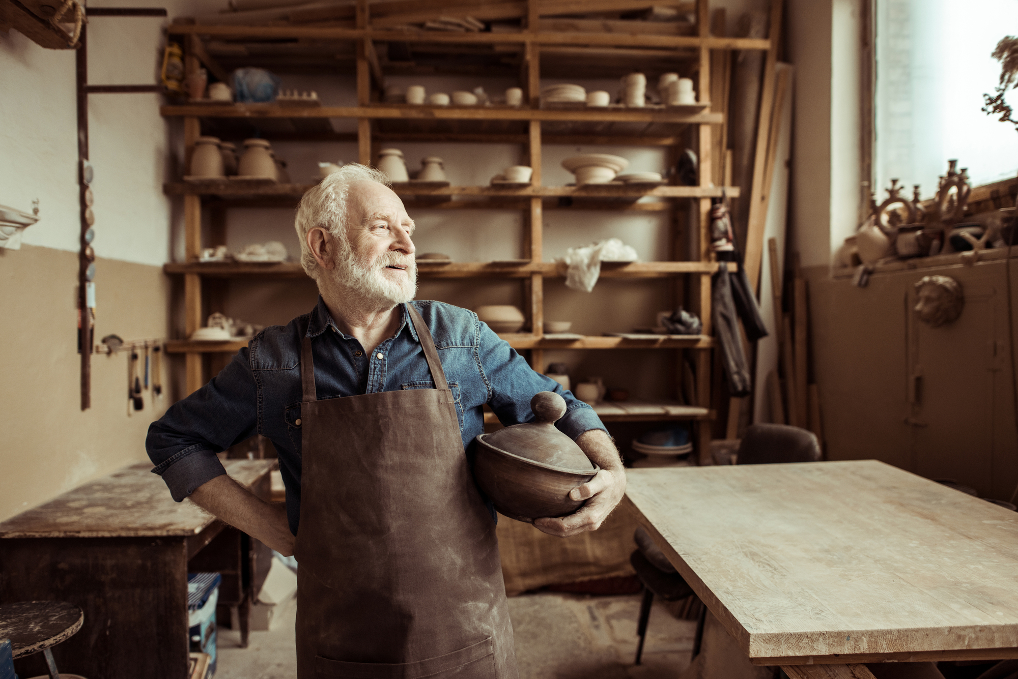 An elderly man with a white beard stands in a pottery studio, wearing a denim shirt and apron. He holds a ceramic pot and looks thoughtfully to the side. Shelves filled with pottery and tools surround him.