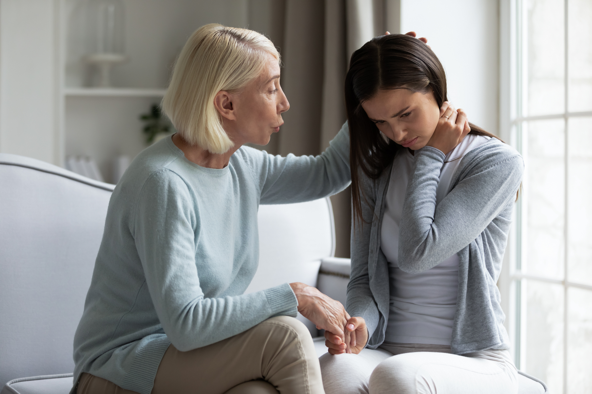 An older woman with short gray hair comforts a younger woman with long brown hair sitting beside her on a couch. The younger woman looks distressed, with her head down and hand on her neck, while the older woman offers support.