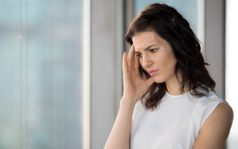 A worried woman with shoulder-length dark hair stands by a window, touching her temple with a thoughtful expression. She wears a white sleeveless top, and the background shows a blurred cityscape.