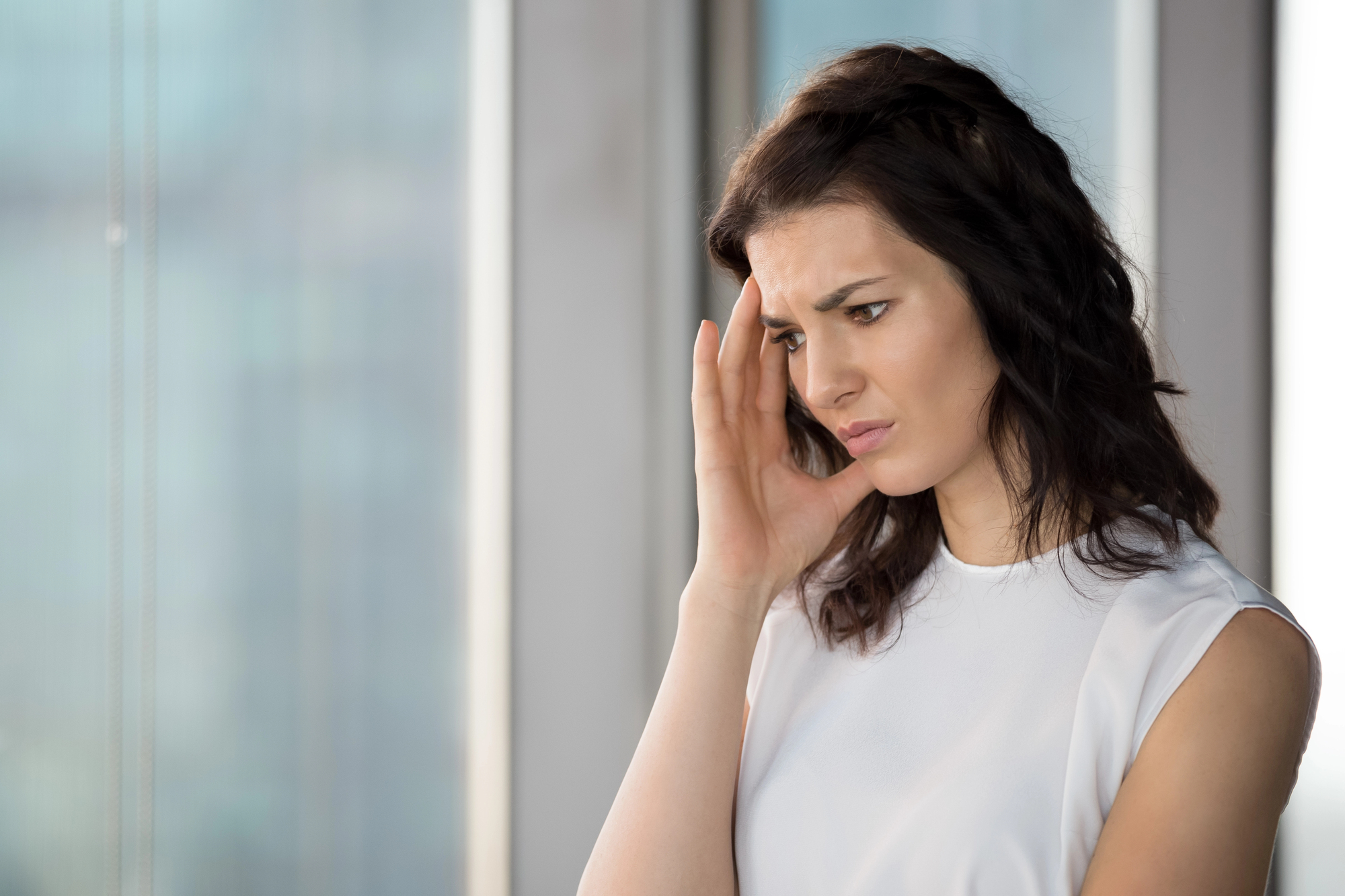 A worried woman with shoulder-length dark hair stands by a window, touching her temple with a thoughtful expression. She wears a white sleeveless top, and the background shows a blurred cityscape.