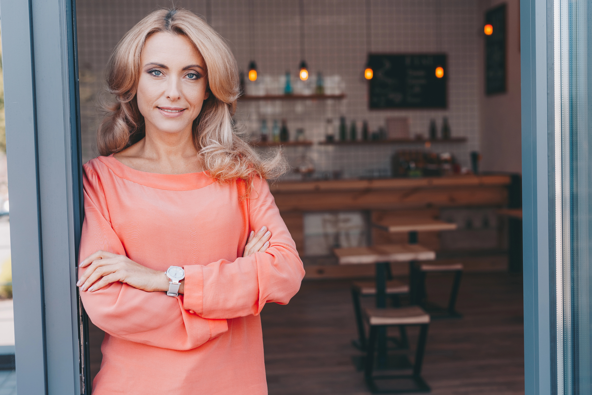 A woman with long, wavy hair stands in a doorway, smiling and crossing her arms. She's wearing an orange blouse and a watch. The background shows a cozy cafe setting with wooden tables and dim lighting.