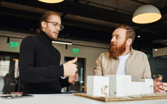 Two men are engaged in a discussion around an architectural model on a table. One man gestures with his hand while the other listens attentively. The setting appears to be a modern office with overhead lights.