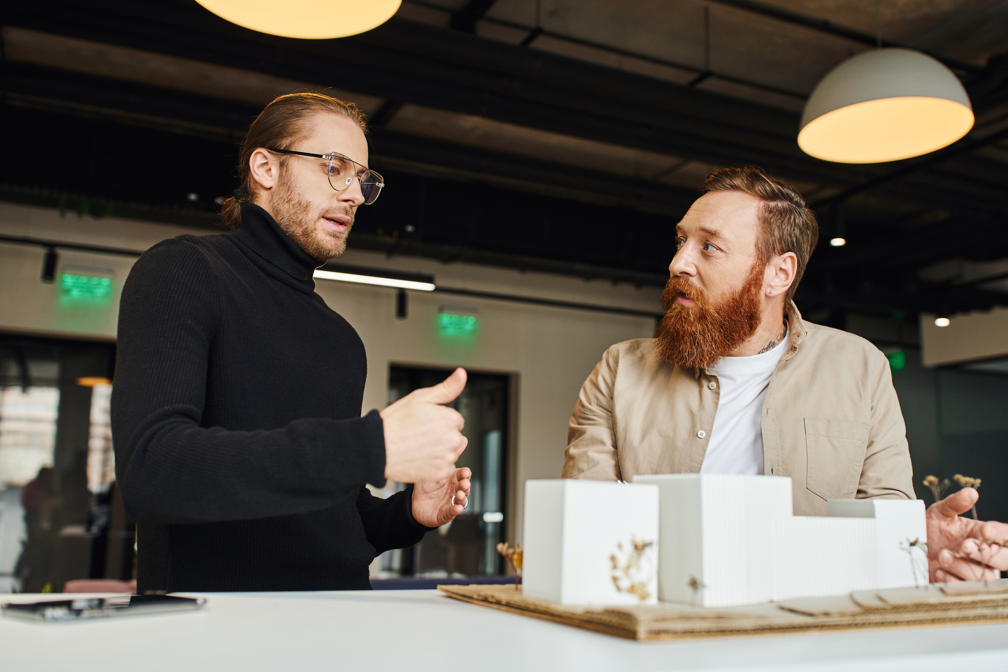 Two men are engaged in a discussion around an architectural model on a table. One man gestures with his hand while the other listens attentively. The setting appears to be a modern office with overhead lights.