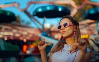 A woman wearing pink sunglasses and stylish earrings poses confidently with thumbs pointing towards herself. She is standing outdoors in front of a blurred amusement park ride. The sunlight creates a vibrant and lively atmosphere.