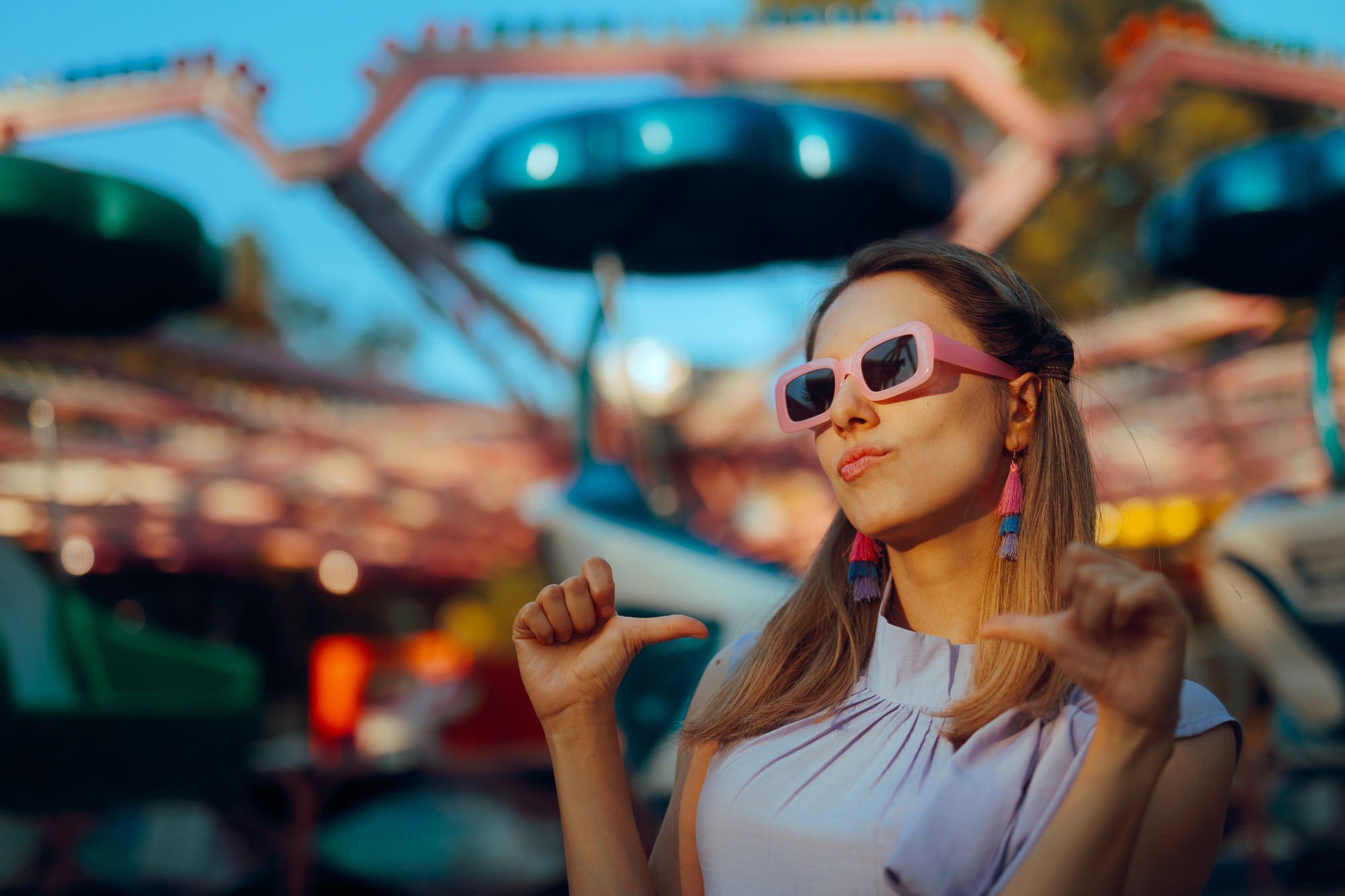 A woman wearing pink sunglasses and stylish earrings poses confidently with thumbs pointing towards herself. She is standing outdoors in front of a blurred amusement park ride. The sunlight creates a vibrant and lively atmosphere.