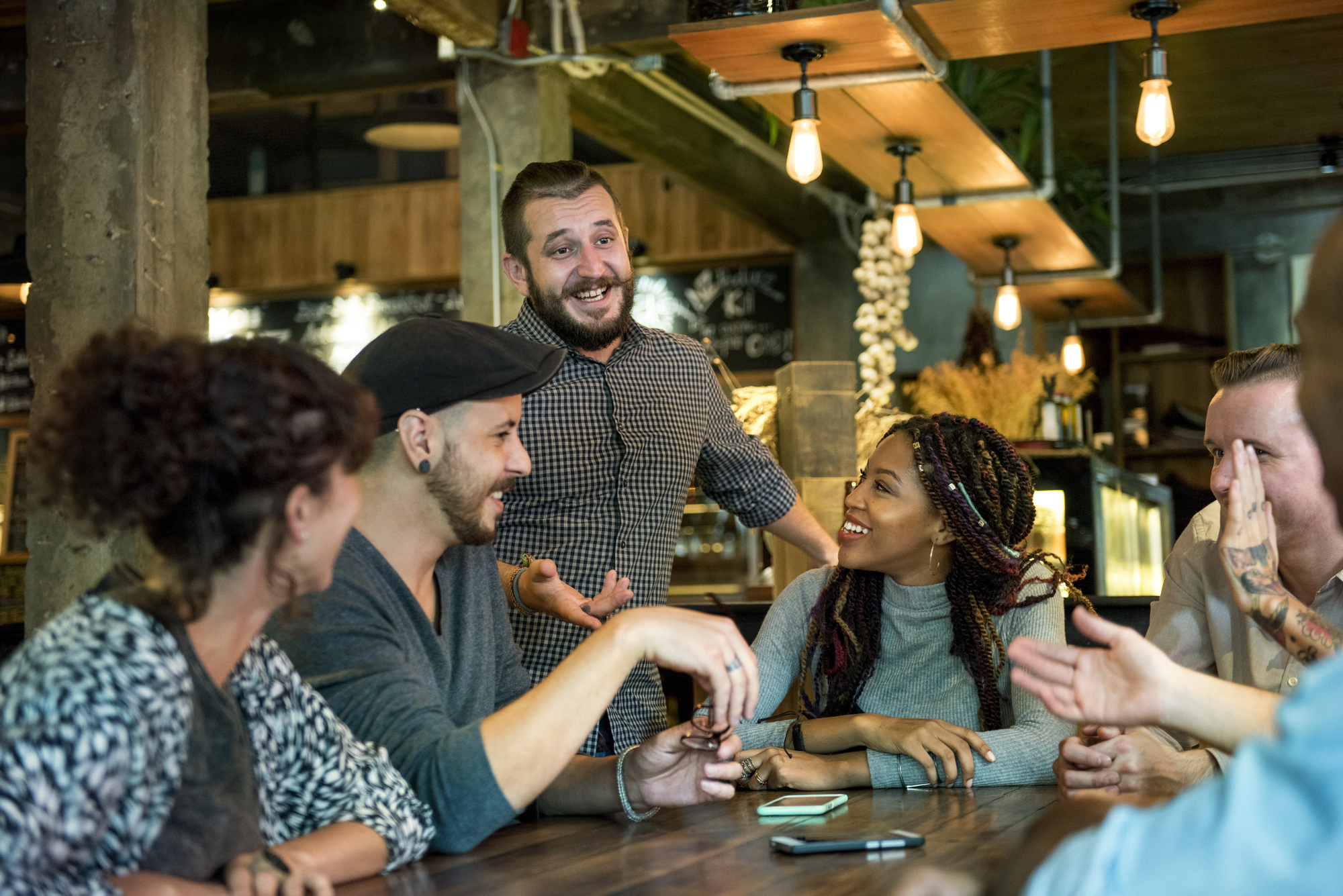 A group of six people sitting around a table in a warmly lit cafe, engaged in lively conversation and laughter. One person is standing, smiling, and talking animatedly while the others listen and respond with smiles and gestures.