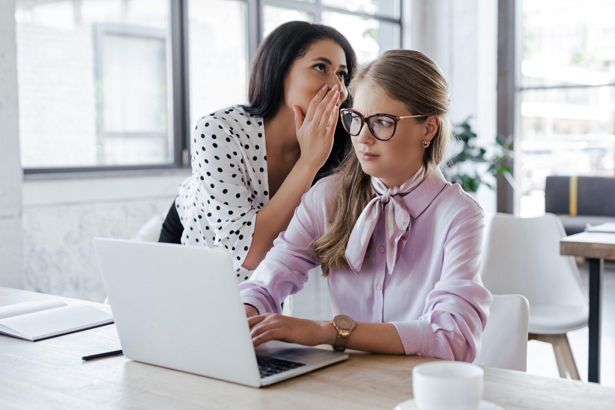 A woman in a polka dot blouse whispers to a woman in a pink blouse who is sitting at a desk with a laptop. They are in a bright office environment with large windows in the background.